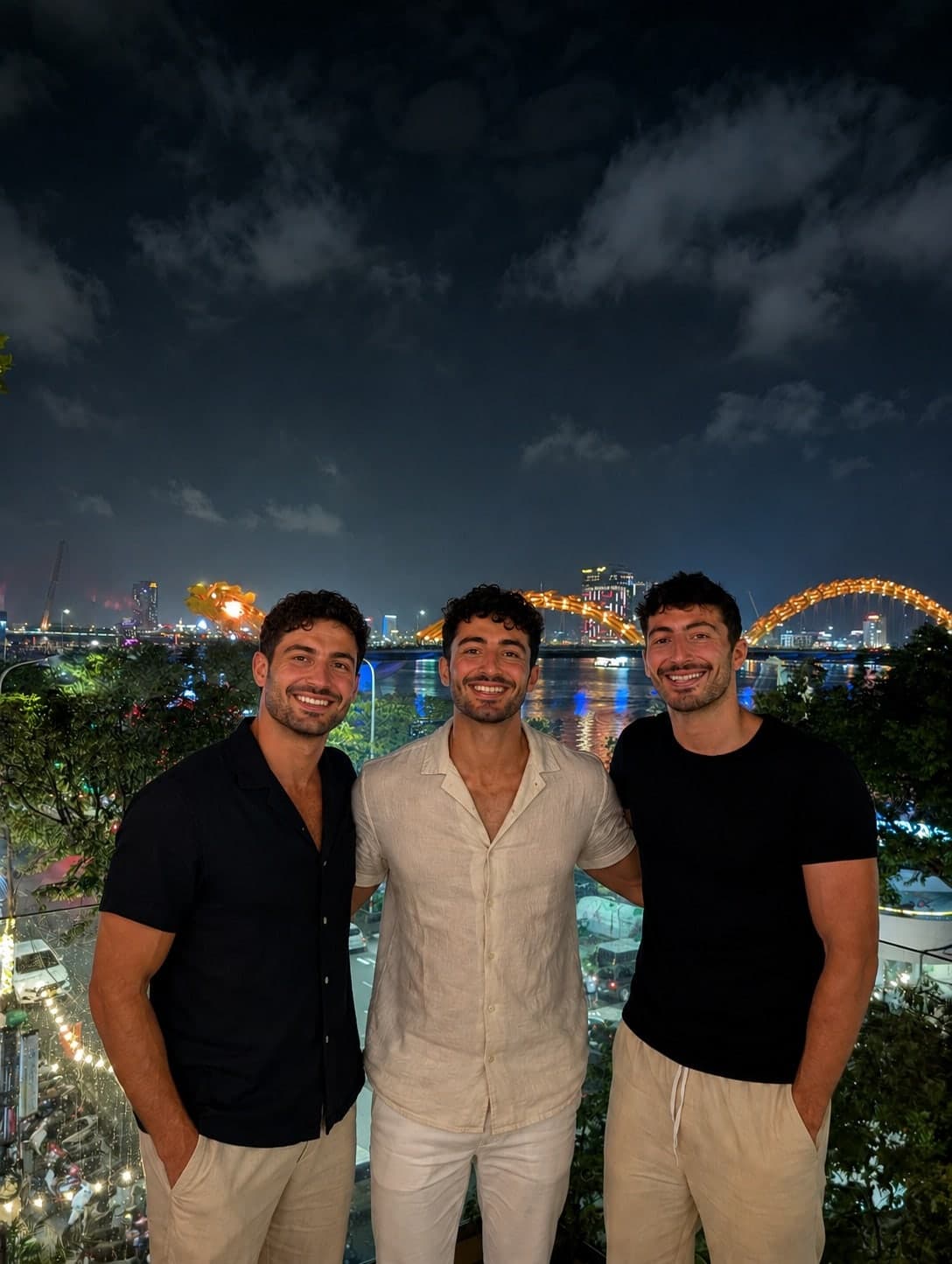 The three Nxeon co-founders on a rooftop in Da Nang, Vietnam, with the Dragon Bridge lit up behind them at night